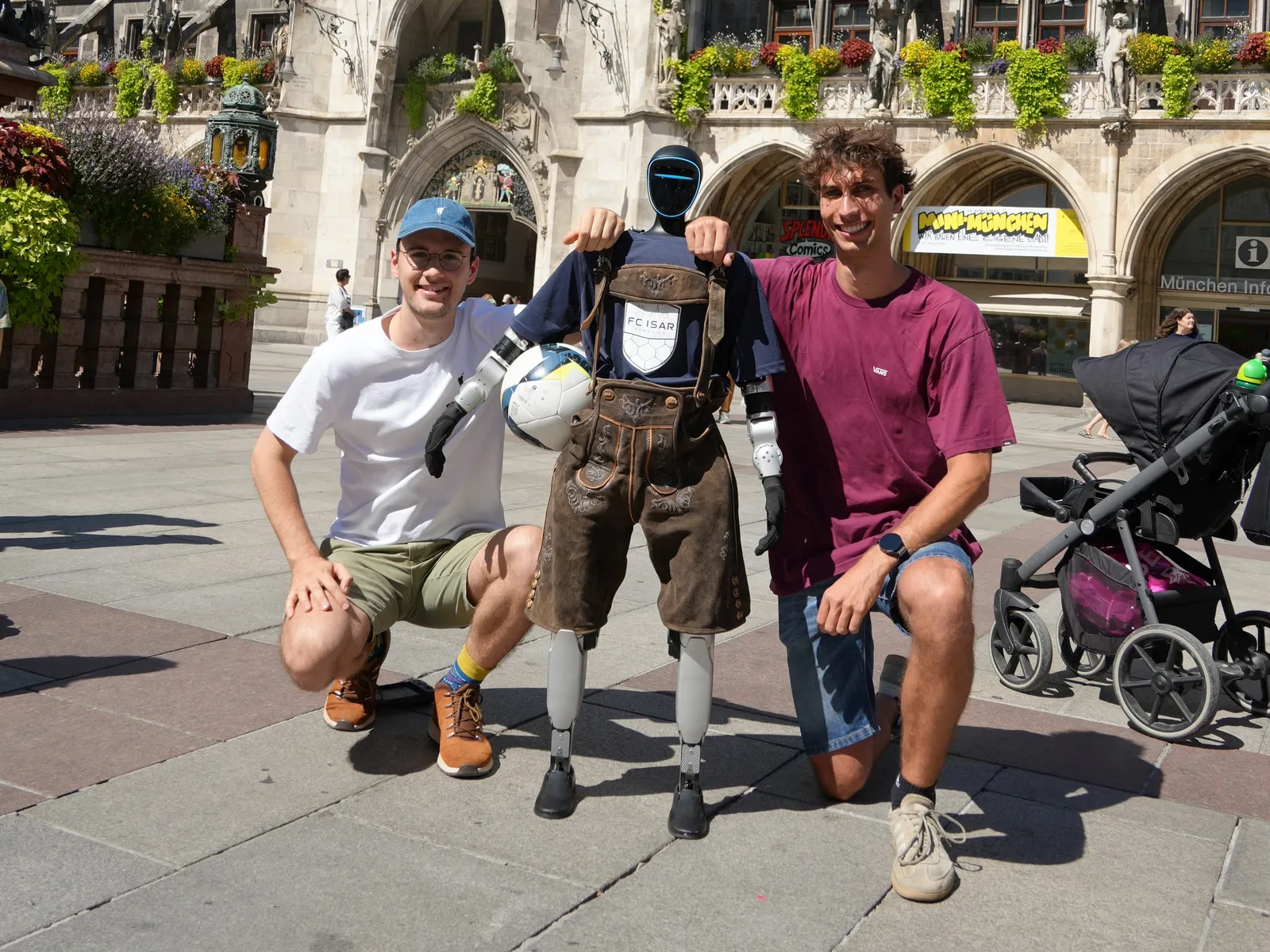 Two team members kneeling either side of the humanoid robot in Lederhosen, in front of Munich's New Town Hall